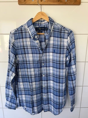 Blårutig skjorta från Ralph Lauren - Klassisk blårutig skjorta från Ralph Lauren med button-down krage och broderad logga på bröstet. Skjortan har långa ärmar, knappar framtill och är tillverkad i mjuk bomull. Perfekt för en avslappnad men ändå stilren look.