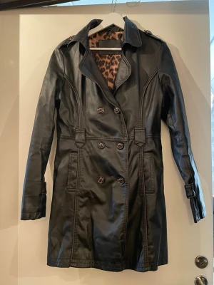 Svart lång skinnjacka med leopardfoder - Snygg svart lång skinnjacka i trenchcoat-modell med dubbelknäppning och markerad midja. Insidan har ett coolt leopardmönstrat foder som ger extra edge. Jackan har axelklaffar och fickor framtill. Den har repor o slitage på viss ställen. Det står L men passar bra på storlek M också.