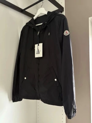 Svart vindjacka från Moncler - Snygg svart vindjacka från Moncler med dragkedja, huva med dragsko och två fickor framtill. Jackans qr och blipp funktionen funkar som hänvisar till monclers hemsida. Storlek 2 vilket motsvarar M-S