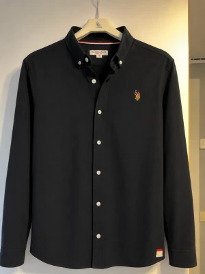 polo ralph lauren Mörkblå skjorta  - Klassisk mörkblå skjorta från polo ralph lauren med broderad logga på bröstet. Skjortan har knappar framtill, button down-krage och långa ärmar. Tillverkad i mjuk bomull som känns skön mot huden. Perfekt för en stilren och avslappnad look.