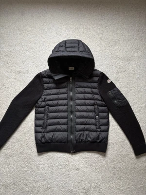 Svart cardigan från Moncler - Snygg svart cardigan från Moncler med quiltad front och huva samt stickade ärmar och rygg. Jackan har dragkedja framtill, två sidofickor med knappar och en extra ficka på ärmen med Moncler-logga. Perfekt för kalla vinterdagar och riktigt trendig look.