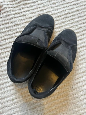 Axel arigato skor  - Säljer ett par svarta slip-on sneakers med breda kardborreband. Skorna har en låg profil och ovandel i mocka som ger en stilren och enkel look. Sulan är vit och kontrasterar snyggt mot det svarta. Perfekta för dig som gillar minimalistisk stil.