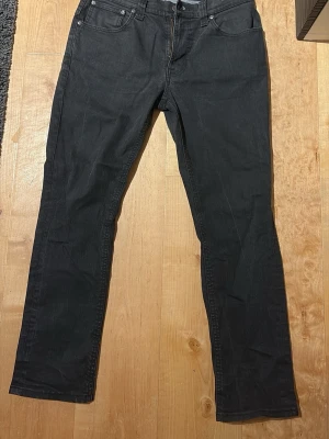 Svarta jeans från Nudie  - Säljer ett par svarta jeans från Nudie med klassisk femficksmodell och raka ben. Jeansen har bälteshällor och dragkedjegylf. Materialet är denim i bomull med en mjuk känsla. Perfekta för en avslappnad och stilren look. Storlek W33 L32