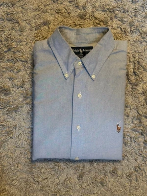 Ljusblå skjorta från Ralph Lauren - Klassisk ljusblå skjorta från Ralph Lauren med button-down krage och broderad logga på bröstet. Skjortan har långa ärmar och vita knappar framtill. Perfekt till en stilren och avslappnad look.