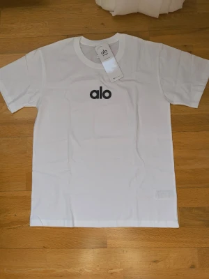 Vit t-shirt från Alo Yoga - Snygg vit t-shirt från Alo Yoga med svart logga tryckt på bröstet. Klassisk rund halsringning och korta ärmar. Tillverkad i mjuk bomull som känns skön mot huden. Perfekt till ett avslappnat streetwear-look.