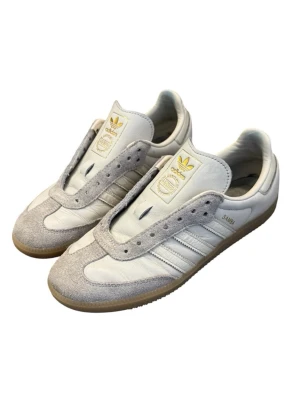 Adidas Samba - Utan skosnören Klassiska Adidas Samba sneakers i vitt läder med ljusgrå mockadetaljer och gummisula. Ikonisk design med tre vita ränder på sidan och guldfärgad Adidas-logga på tungan. Perfekt för dig som gillar retrostil och tidlösa sneakers.
