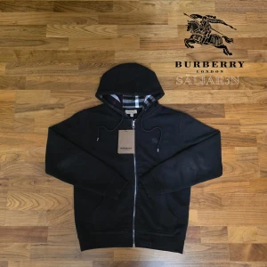 Svart Burberry Hoodie  - Letar du efter en stilren och lyxig hoodie? Denna helt nya grå Burberry-hoodie kombinerar elegans med hög komfort. Den har Burberrys ikoniska rutmönster i fodret och en diskret logga på bröstet för en exklusiv touch. Tillverkad av mjukt, slitstarkt material och utrustad med dragkedja samt huva – både praktisk och stilfull.  📌 Skick: Ny med taggar. 📦 Snabb leverans – skickas samma dag (om ej hindr)