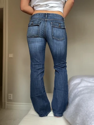 Blå bootcut jeans med ficklock - Snygga blå bootcut jeans med ficklock bak och låg midja. Jeansen har klassisk femficksdesign och är tillverkade i ett stretchigt denimtyg som sitter skönt. Perfekta för en avslappnad och trendig look.