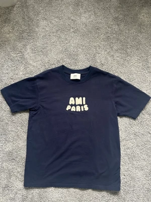 Mörkblå t-shirt från Ami Paris - Snygg mörkblå t-shirt från Ami Paris med ett vitt tryck på bröstet där det står 'AMI PARIS' i bubbliga bokstäver. Klassisk passform och rund halsringning. Perfekt till jeans eller shorts för en clean look.
