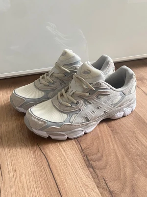 Beige ASICS sneakers med mesh och mocka - Snygga sneakers från ASICS i beige och ljusgrå toner. Ovandelen är i mesh och mocka för en sportig och trendig look. Skorna har klassisk snörning och en chunky sula som ger extra komfort. Perfekta för dig som gillar stilrena och neutrala färger.