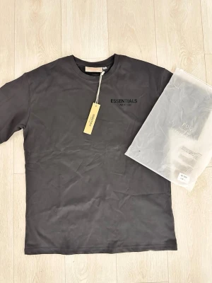 Svart Essentials Fear of God t-shirt - Säljer en svart t-shirt från Essentials Fear of God med diskret logga på bröstet. T-shirten har klassisk rund hals och korta ärmar, perfekt för en clean och stilren look. Materialet är mjukt och känns skönt mot huden.