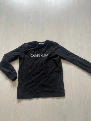Svart sweatshirt från Calvin Klein - Svart sweatshirt från Calvin Klein med vit logga tryckt på bröstet och en liten 'JEANS'-patch på ryggen. Tröjan har rund halsringning, långa ärmar och är gjord i mjukt bomullsmaterial. Perfekt för en avslappnad och stilren look.