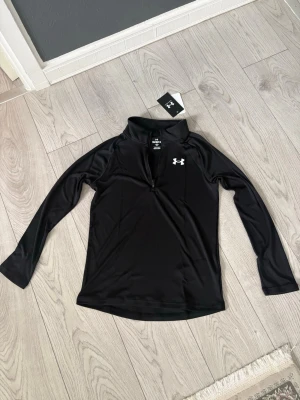 Svart träningströja från Under Armour - Snygg svart långärmad träningströja från Under. Tillverkad i ett stretchigt funktionsmaterial som andas, perfekt för löpning eller gym. Tight passform och hög krage för extra komfort. Den var tyvärr för liten för mig den är helt ny med prislapp på