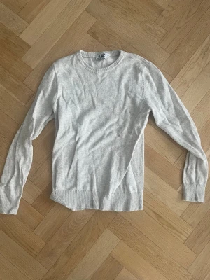 Ljusgrå stickad tröja - En stilren ljusgrå stickad tröja med rund halsringning och långa ärmar. Tröjan har ribbade muddar vid ärmslut och nederkant, vilket ger en klassisk look. Perfekt att matcha med jeans eller shorts för en enkel och snygg outfit. Köpt från stilgruvan som är Oliver kastells.