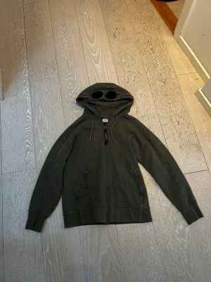 Mörkgrön hoodie från C.P. Company - Just nu ligger denna ikoniska cp company hoodien ute för försäljning. Den har de unika glassögonen på luvan. Men har tyvärr fått några målarfärgs fläckar som inte går att ta bort.
