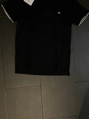 Svart piké från Selected Homme - Klassisk svart pikétröja från Selected Homme med vit broderad logga på bröstet och vita ränder på krage och ärmslut. Tröjan har korta ärmar och knäppning vid halsen. Perfekt till en clean och stilren look.