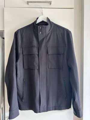 J Lindeberg Field Jacket - Snygg svart jacka med ståkrage och två stora bröstfickor med lock samt två dragkedjeförsedda sidofickor. Jackan har minimalistisk design och stängs med dragkedja och tryckknappar. Perfekt för dig som gillar stilrena plagg.