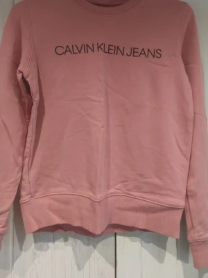 Rosa sweatshirt från Calvin Klein Jeans - Säljer en rosa sweatshirt från Calvin Klein Jeans med svart logotyptryck på bröstet. Tröjan har rund halsringning, långa ärmar och ribbade muddar vid ärmslut och nederkant. Perfekt för en avslappnad och trendig stil.