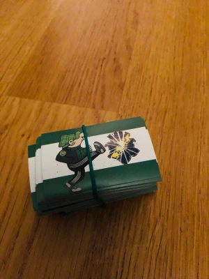 Samlarbilder med tecknad figur - Ett knippe samlarbilder med grönvit baksida, hålls ihop med ett gummiband. Motivet visar en tecknad figur i gröna kläder som sparkar mot ett emblem. Perfekt för dig som gillar samlarkort eller vill ha något unikt till din bokhylla.