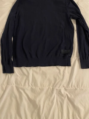 Mörkblå stickad tröja Tommy Hilfiger - Snygg mörkblå stickad tröja från Tommy Hilfiger i storlek L. Tröjan har rund halsringning, ribbade muddar vid ärmslut och nederkant samt en diskret logolapp vid nederkanten. Perfekt till lager på lager och enkel att matcha.