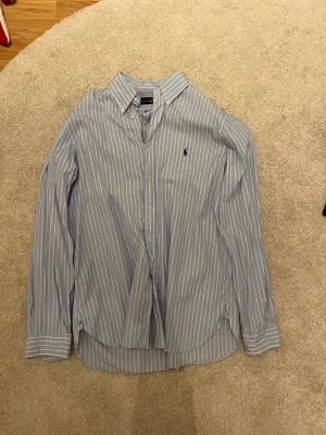 Blårandig skjorta från Ralph Lauren - Klassisk blårandig skjorta från Ralph Lauren med vit knappslå och button-down krage. Skjortan har lång ärm och den ikoniska broderade loggan på bröstet. Tillverkad i mjuk bomull som känns skön mot huden. Perfekt för en stilren och avslappnad look.