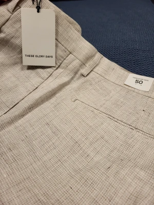Beige linnebyxor These Glory Days 50 M - Stilrena beige linnebyxor från These Glory Days med rak passform och diskret struktur i tyget. Byxorna har hällor för bälte, fickor bak och klassisk knappstängning. Perfekta för en avslappnad men snygg look.