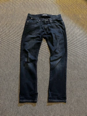 Acne jeans - Säljer ett par svarta jeans från Nudie med klassisk femficksmodell och rak passform. Jeansen har en snygg slitning på vänster knä och subtila slitningar vid fickor och benslut. Tillverkade i denim med lite tvättad look.