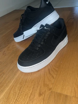 Nike Air Force 1 svart/vit sneakers - Svarta Nike Air Force 1 sneakers med vit sula och klassisk swoosh-logga på sidan. Skorna har lågt skaft, perforerad tå och är tillverkade i mocka. Snygg kontrast mellan svart ovandel och vit yttersula. Perfekta för en stilren streetlook. Väldigt sällsynta och kommer i vågor och blir ibland väldigt svåra att hitta