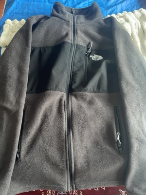 Grå fleecejacka från The North Face - Mysig grå fleecejacka från The North Face med svarta detaljer och dragkedjor. Jackan har hög krage, tre fickor med dragkedja och logga på bröstet. Perfekt för kyliga dagar och enkel att matcha med olika outfits.