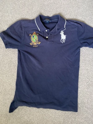 Marinblå piké från Polo Ralph Lauren - Snygg marinblå pikétröja från Polo Ralph Lauren med vit broderad logga på bröstet och emblem. Klassisk krage med knappar och vit piping längs kragen. Tillverkad i mjuk bomull och har korta ärmar.