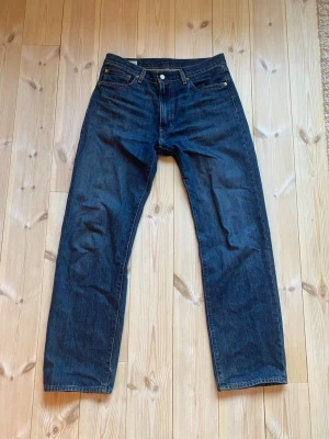 Mörkblå raka jeans från Levi's - Säljer ett par klassiska mörkblå jeans från Levi's med rak passform och fem fickor. Jeansen har en tidlös look med knappgylf och Levi's ikoniska läderpatch bak i midjan. Tillverkade i slitstark denim som passar perfekt till alla tillfällen.