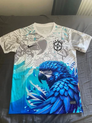 Fotbolls T-shirt brazil  - Unik sporttröja från Nike med Brasilien CBF-logga. T-shirten har ett färgstarkt tryck med en stor blå papegoja och detaljer i vitt, grått och turkost. Tillverkad i Dri-FIT-material som andas, perfekt för träning eller streetwear. Storlek S