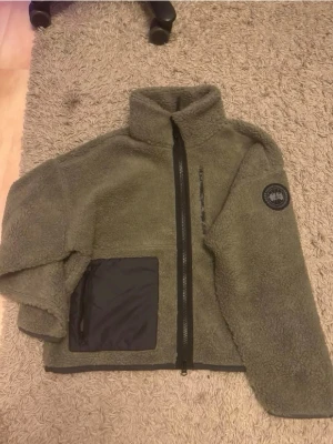 Olivgrön teddyjacka från Canada Goose - Säljer en olivgrön teddyjacka från Canada Goose med hög krage, hel dragkedja och flera fickor med svarta detaljer. Jackan har ett patch på ärmen och är tillverkad i mjukt, fluffigt material som håller dig varm och bekväm.