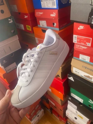 Vita Adidas sneakers med guldlogga - Fräscha vita sneakers från Adidas med klassiska tre ränder på sidan och gulddetaljer på tungan och loggan. Skorna har en låg profil och är gjorda i syntetmaterial med snörning. Perfekta för en clean och stilren look.