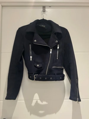  biker skinnjacka med bälte - Snygg svart biker skinnjacka från Zara med silvriga dragkedjor, fickor och ett justerbart bälte i midjan. Jackan har klassisk krage och långa ärmar med dragkedjor vid ärmsluten. Perfekt för en edgy look.