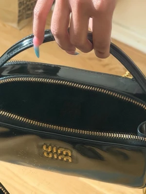 Svart lackväska  - Snygg svart handväska i lack från  med guldiga detaljer och dragkedjor. Väskan har ett stilrent, rektangulärt format och ett handtag upptill. Perfekt för dig som gillar en trendig och lyxig look. Väskband medföljer 