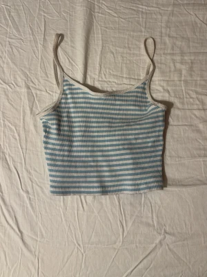 Blåvit ribbad cropped linne - Säljer ett blåvitt ribbat linne med smala axelband och croppad passform. Linnet har tunna horisontella ränder och vita kanter runt halsringning och axelband. Perfekt för sommaren och enkel att matcha med jeans eller shorts.