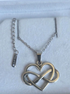 Silverfärgat halsband med hjärthänge - Snyggt halsband i silverfärgat rostfritt stål med ett hänge i form av ett hjärta och en oändlighetssymbol som är sammanflätade. Kedjan är tunn och har en förlängningsdel för justerbar längd. Perfekt accessoar för att lyfta din outfit.