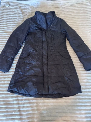 Svart lång dunjacka med bälte - Snygg svart lång dunjacka med markerad midja och bälte bak. Jackan har hög krage, långa ärmar och quiltade sömmar i ett cirkulärt mönster. Perfekt för kalla vinterdagar och ger en stilren siluett.