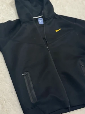 Svart Nike hoodie med gul logga - Snygg svart hoodie från Nike med dragkedja framtill och två fickor med dragkedja. Klassisk gul Nike-logga på bröstet. Hoodien har huva och är tillverkad i ett mjukt material som känns bekvämt. Perfekt för en sportig och avslappnad stil.