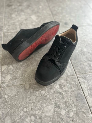 Christina  louboutin - Säljer ett par svarta sneakers med lågt skaft och klassisk snörning. Skorna har en ovandel i tyg och en cool röd yttersula som sticker ut. Insidan är beige och de har en enkel, stilren design som passar till det mesta.