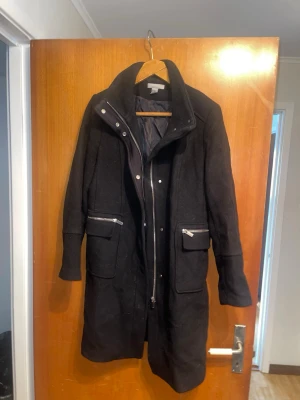 Svart ullkappa med dragkedja - Stilren svart kappa från H&M i ullblandning. Kappan har hög krage, långa ärmar och praktiska fickor med dragkedja framtill. Perfekt för kyliga dagar och ger en snygg siluett med sin klassiska design.