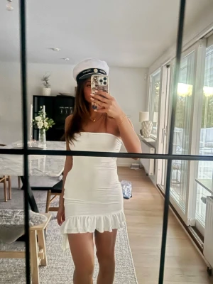 Vit kort klänning med volang - Säljer en snygg vit kort klänning med volangkant och öppen rygg med snörning. Klänningen är axelbandslös och har en tight passform som framhäver figuren. Perfekt för student eller sommarens alla tillfällen! Den är från oh polly och säljs inte längre. 💓💓💓 inte genomskinlig alls och har en skurning vid midjan 