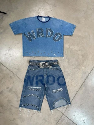 Blå croppad denim-topp med nitar - Trendig croppad topp i blå denim från SH med stora bokstäver 'WRDO' framtill. Toppen har korta ärmar, råa kanter och är dekorerad med nitar längs nederkanten och runt bokstäverna. Perfekt för en edgy streetstyle-look.