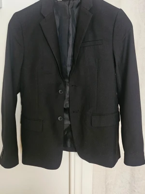 Svart klassisk kavaj med knappar - Stilren svart kavaj med två knappar framtill, klassisk krage och fickor med lock. Perfekt för en dressad look och passar till många olika outfits. Långärmad modell med slät finish och helfodrad insida.