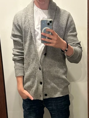 Grå ribbstickad kofta med sjalkrage - Snygg grå ribbstickad kofta med sjalkrage och svarta knappar framtill. Koftan har långa ärmar och en avslappnad passform, perfekt att slänga på över en t-shirt. Materialet ser mjukt och bekvämt ut, och den neutrala färgen gör den enkel att matcha.