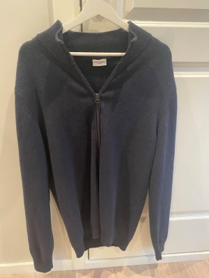 Mörkblå ziptröja från John Henric - Mörkblå stickad ziptröja från John Henric med hög krage och hel dragkedja framtill. Tröjan har lång ärm och ribbade muddar vid ärmslut och nederkant. Perfekt för lager på lager och stilren look.
