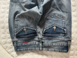 Snygga blå jeans med ficklock  - Säljer ett par blå jeans från Dsquared2 med raka ben och unika bakfickor med ficklock och silverfärgade knappar. Jeansen har klassisk femficksdesign och en liten röd Dsquared2-tag på baksidan. Tillverkade i slitstark denim med snygg tvätt.
