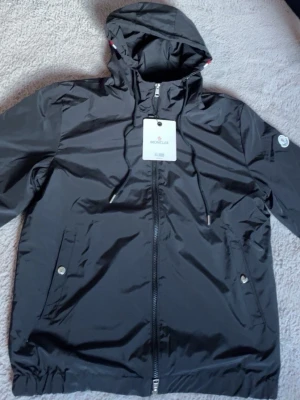 Svart vindjacka från Moncler - Snygg svart vindjacka från Moncler med huva och dragkedja framtill. Jackan har två framfickor med tryckknappar och justerbar dragsko i huvan. Tillverkad i ett lätt, glansigt material och har Moncler-logga på ärmen.