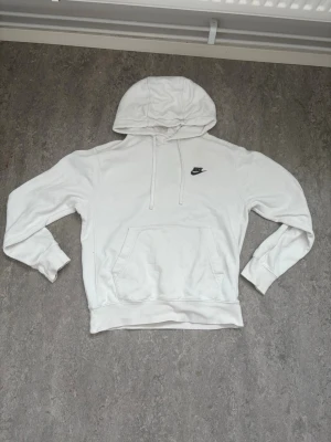 Vit Nike hoodie med huva - Säljer en klassisk vit hoodie från Nike med svart logga på bröstet. Hoodien har en stor huva med dragsnören, ribbade muddar och en praktisk magficka. Materialet är mjukt och bekvämt, perfekt för chill dagar eller när du vill ha en sportig look.